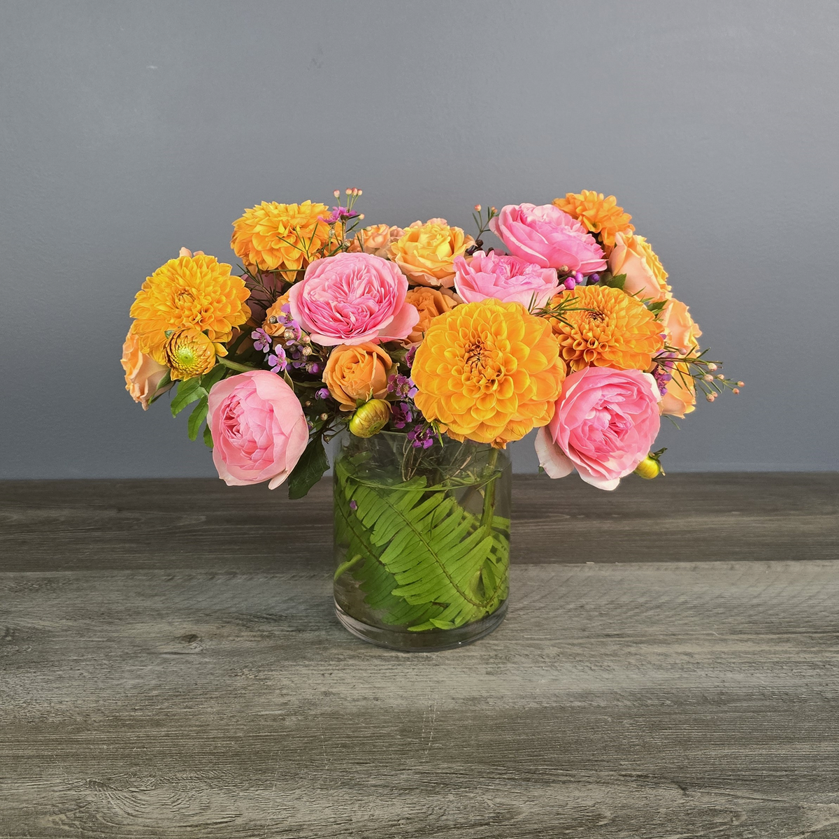 Vibrant mixed bouquet of yellow dahlias, pink garden roses, and peach spray roses arranged in a clear glass vase with green leaf filler.