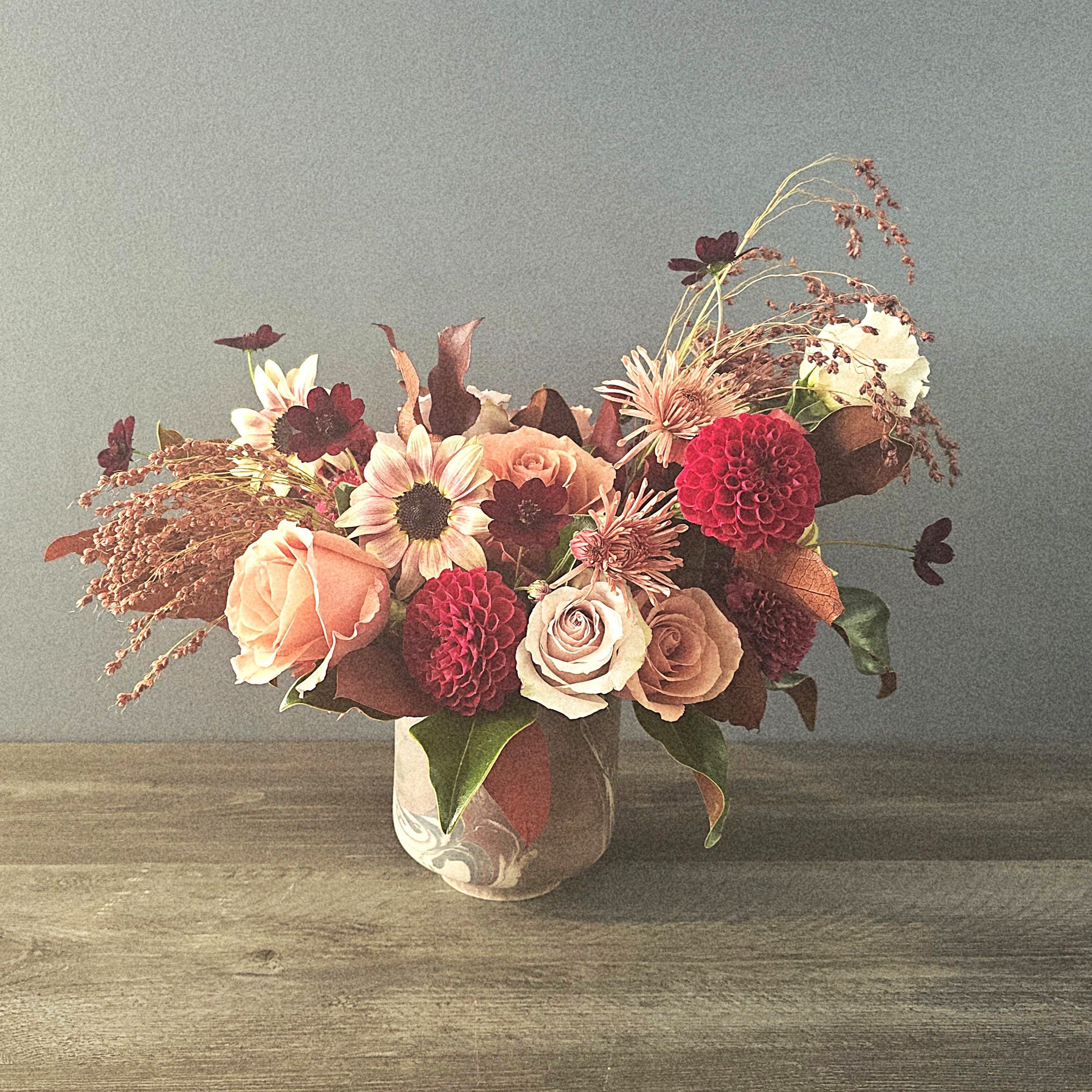 A lush autumn arrangement of burgundy dahlias, cream roses, blush ranunculus, and white daisies in a clear glass vase.