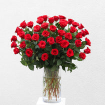 A full, rounded bouquet of vibrant red roses with green stems and foliage arranged in a clear glass vase filled with water.