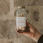 A hand holds a clear glass jar filled with cream-colored safety matches labeled "Safety Matches" against a textured beige wall.