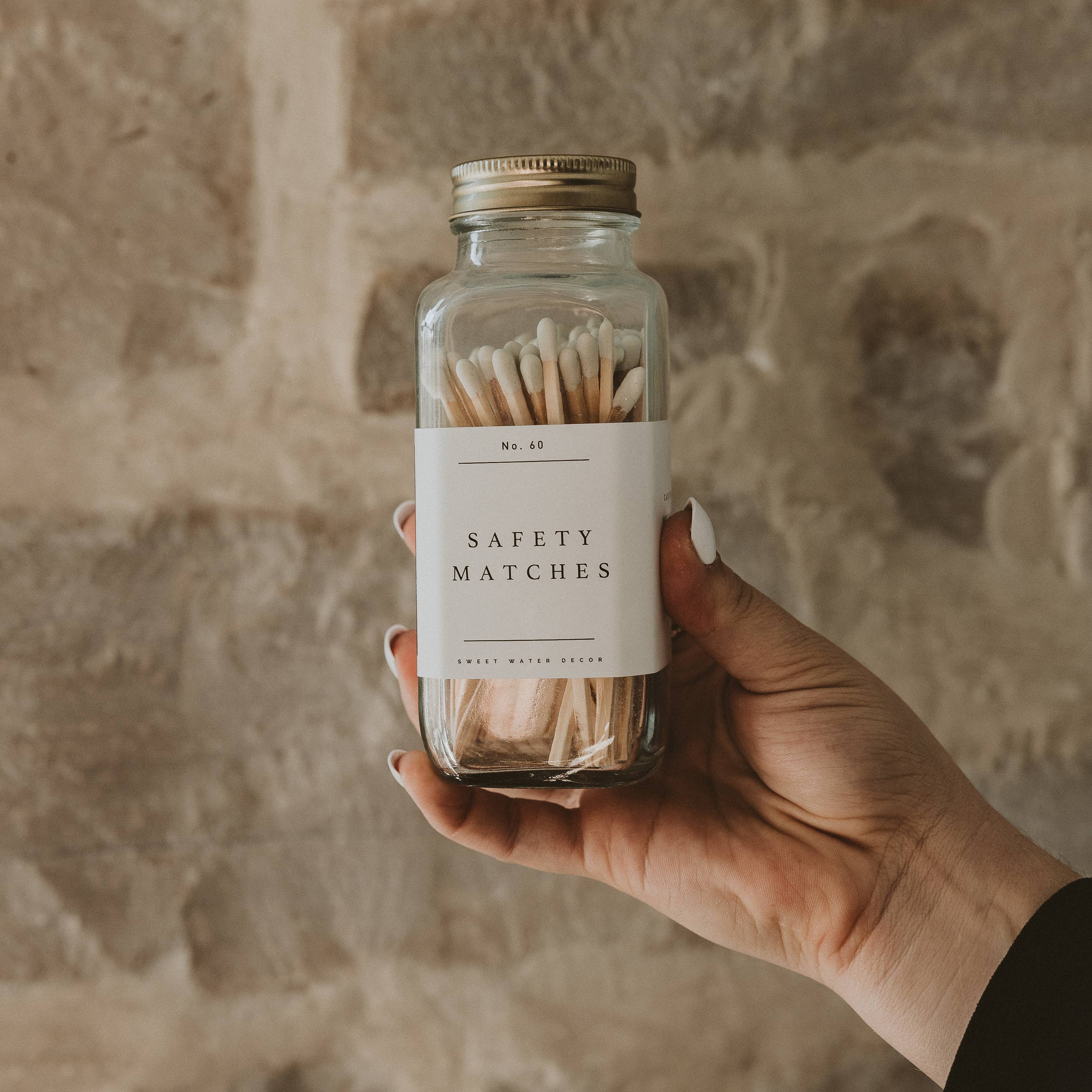 A hand holds a clear glass jar filled with cream-colored safety matches labeled "Safety Matches" against a textured beige wall.