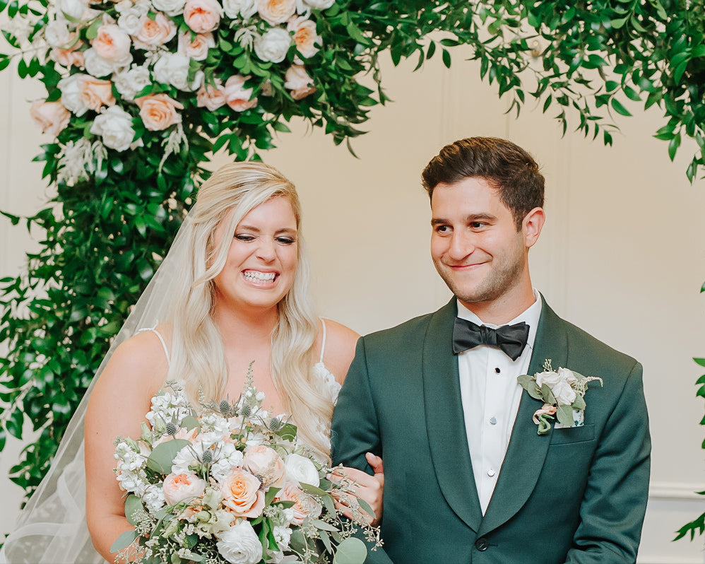 Wedding couple standing under a floral archway with a floral arrangement in the foreground.