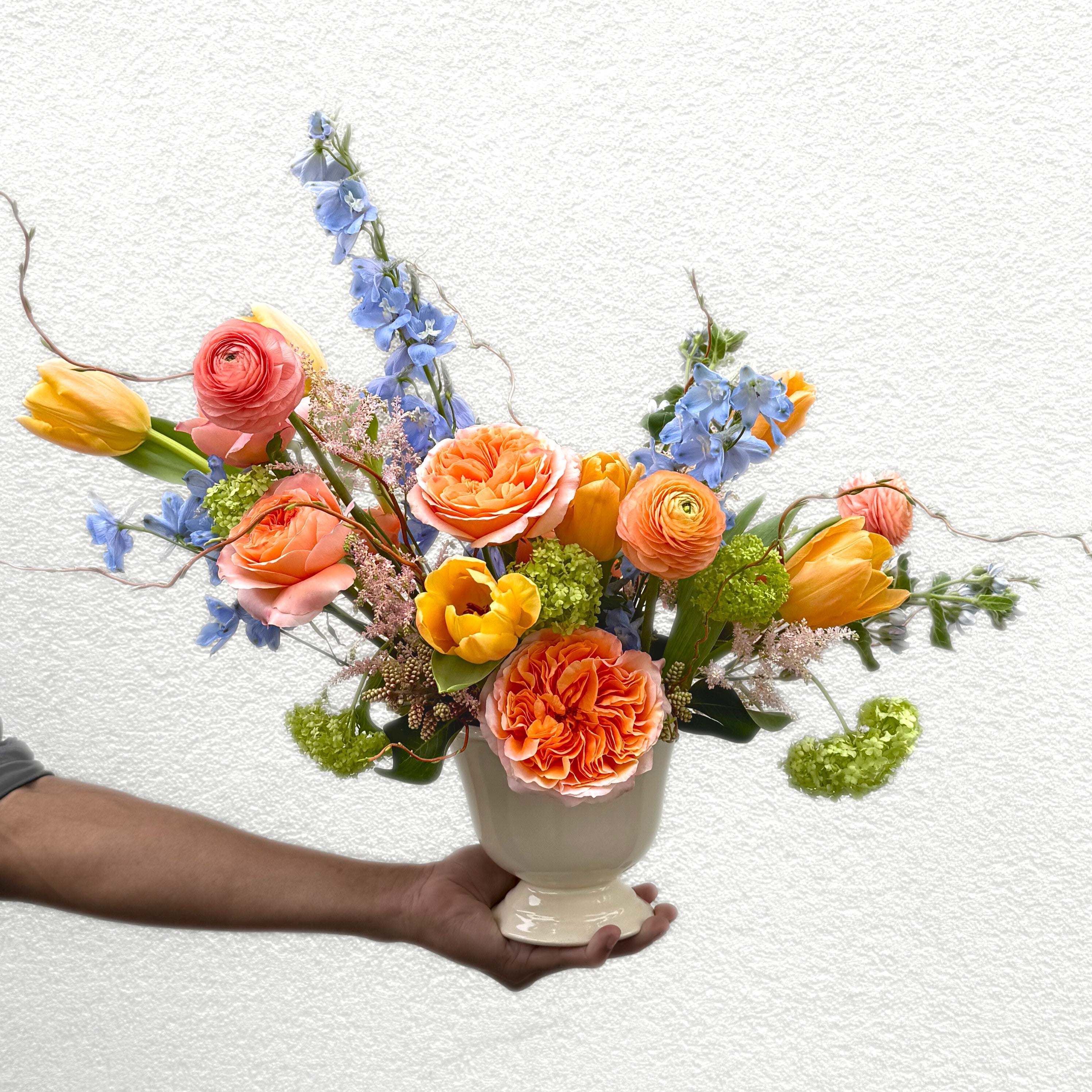 Hand holding a white ceramic vase with coral garden roses, blue delphinium, yellow ranunculus, and green trick dianthus against a white background.