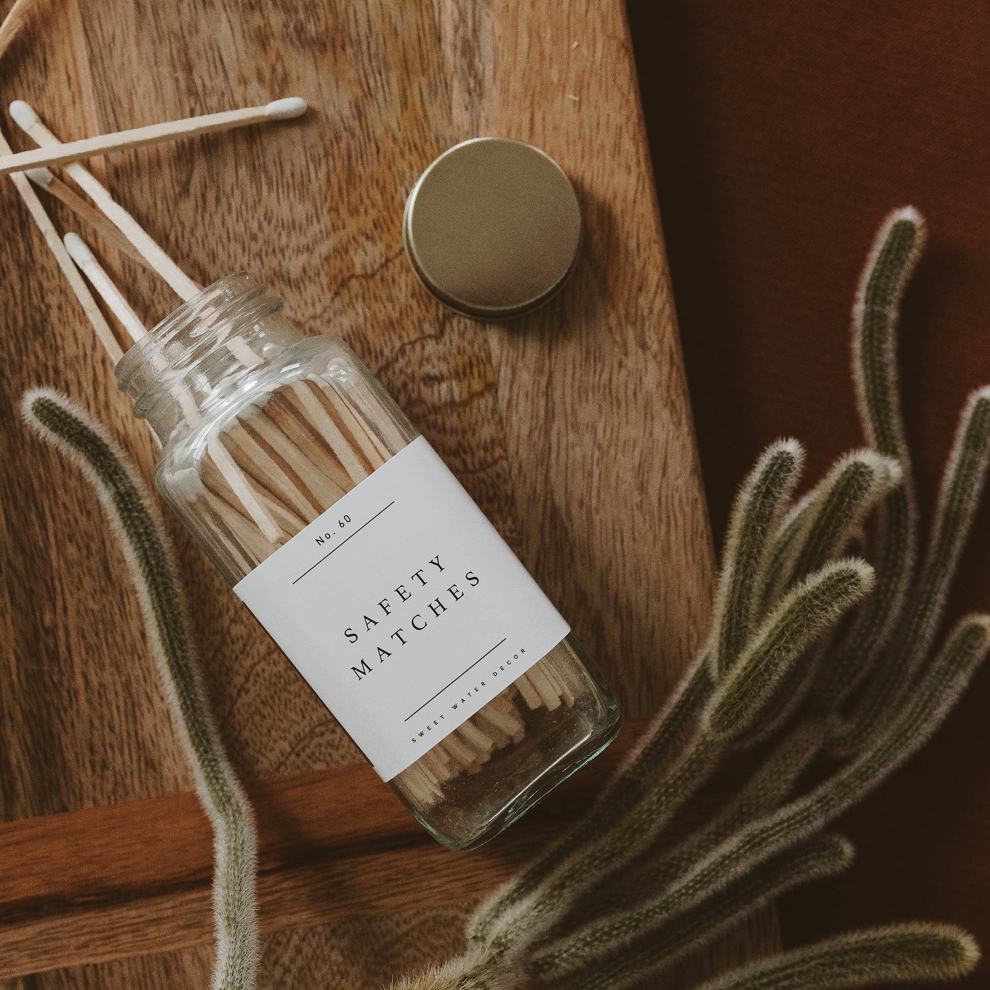 A glass jar of safety matches with a white label sits on a wooden surface next to green dried celosia stems.