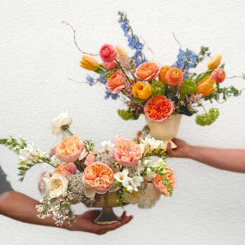 Two hands holding lush bouquets of coral garden roses, orange ranunculus, white daisies, delphinium, and greenery in black and cream vases. — The Flower Bar, Larchmont NY