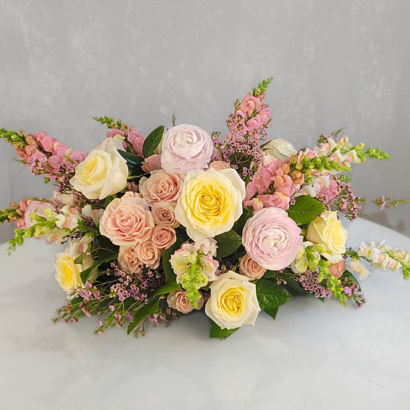 Lush low centerpiece with cream and yellow roses, pink ranunculus, dusty rose spray roses, and burgundy astilbe with green hypericum berries and seasonal gre…