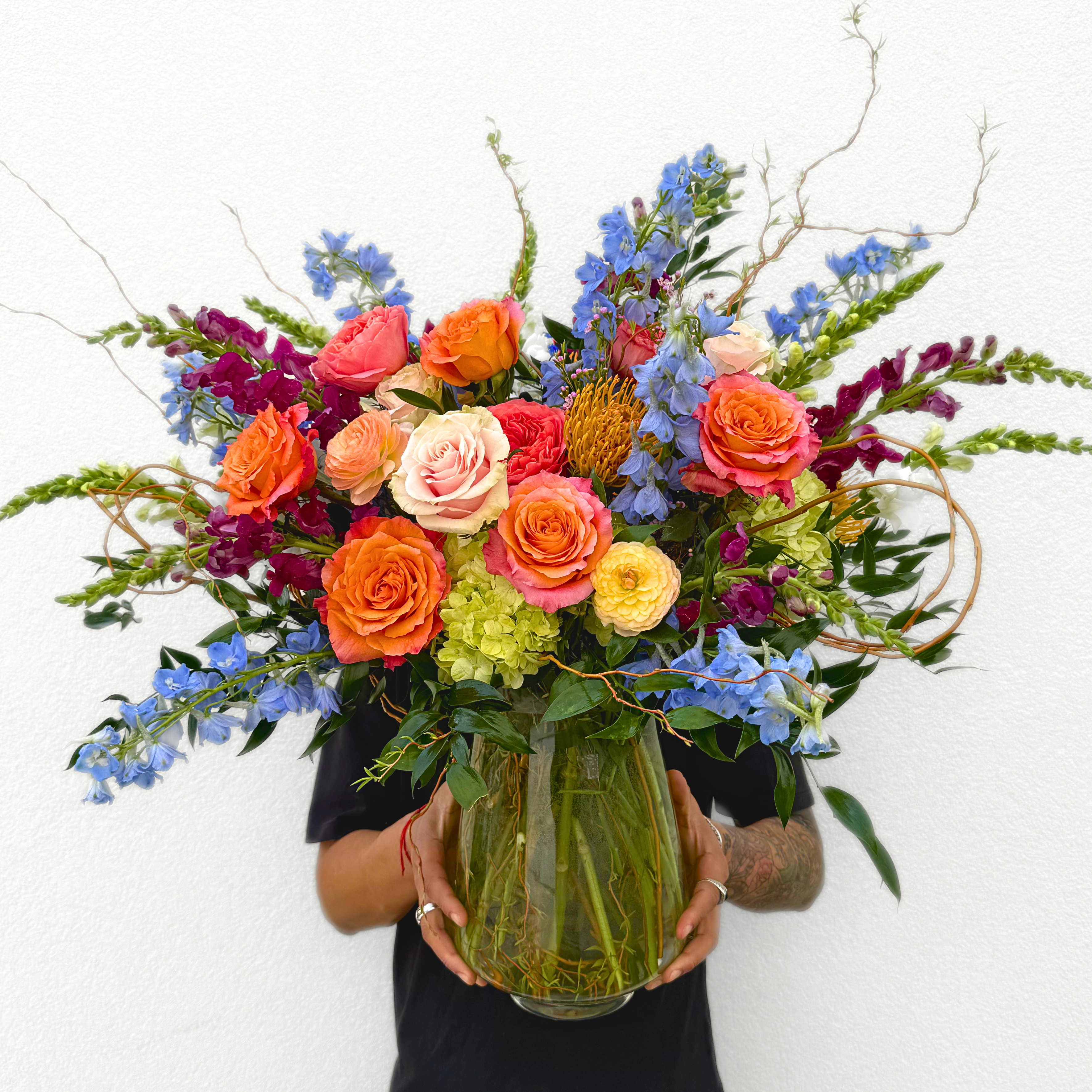 A florist holds a vibrant mixed bouquet in a clear glass vase featuring orange roses, blue delphinium, coral spray roses, burgundy carnations, green hydrange…