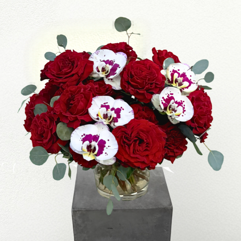 Red roses and white orchids with purple centers arranged in a clear glass vase with eucalyptus greenery on a gray pedestal. — The Flower Bar, Larchmont NY