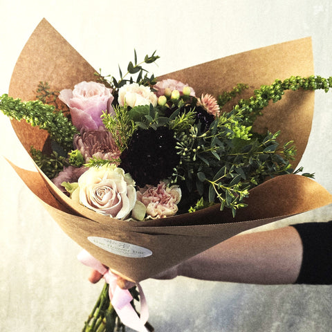 Hand-held bouquet with cream roses, pink carnations, black dahlias, and greenery wrapped in kraft paper. — The Flower Bar, Larchmont NY