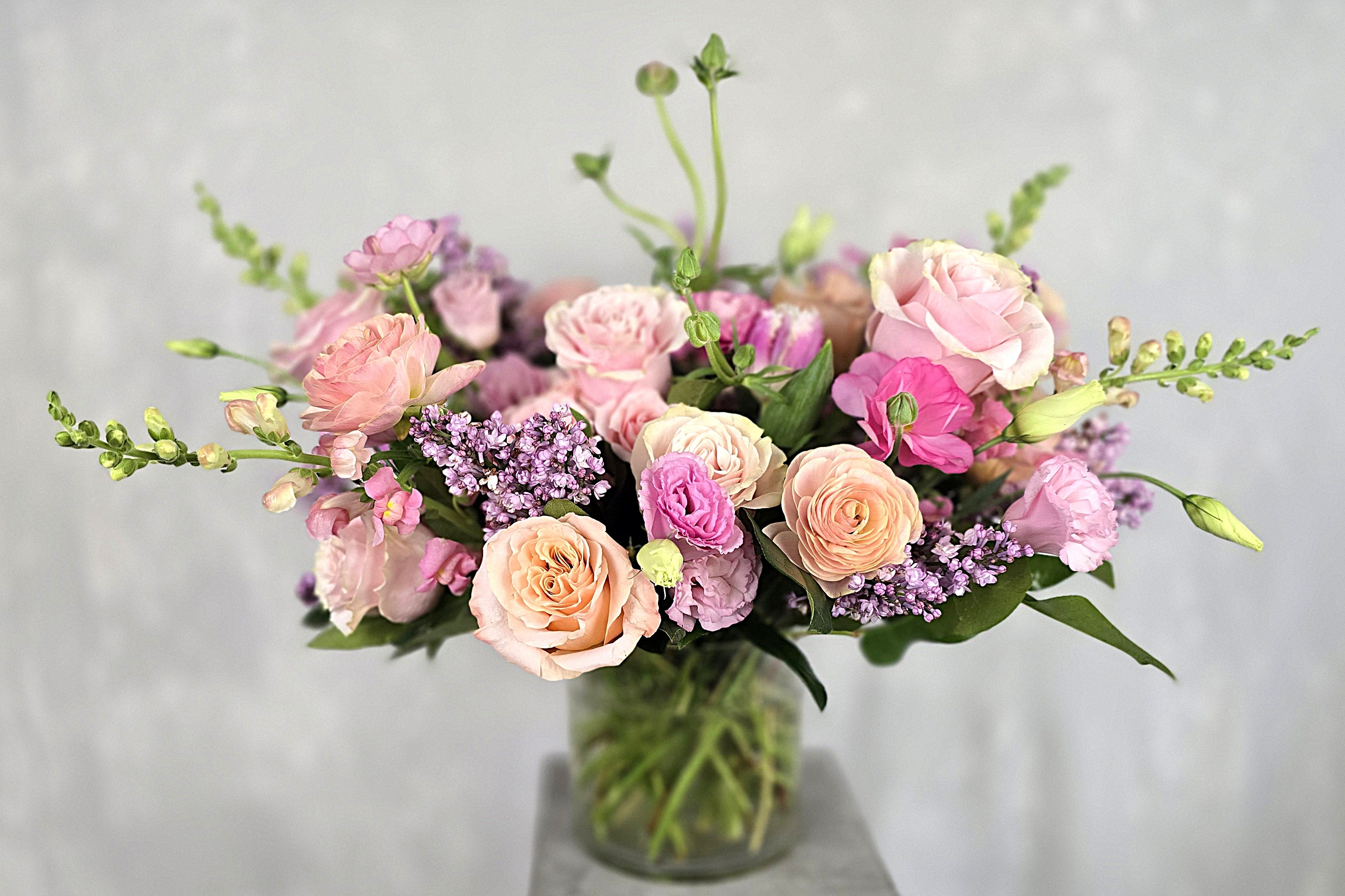 Lush pink and purple floral arrangement in a clear glass vase featuring roses, carnations, lilac, and greenery stems.