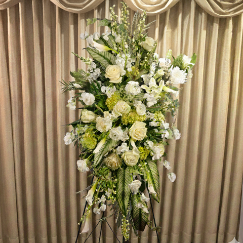 Tall standing funeral spray with cream roses, white carnations, and green foliage against draped beige curtains. — The Flower Bar, Larchmont NY