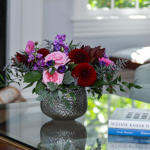 A lush floral arrangement of deep red dahlias, pink roses, purple stock, and magenta carnations in a textured gray vase. — The Flower Bar, Larchmont NY