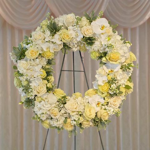 Large circular wreath featuring cream roses, yellow roses, white hydrangeas, and green foliage on a black metal stand against a draped backdrop. — The Flower Bar, Larchmont NY